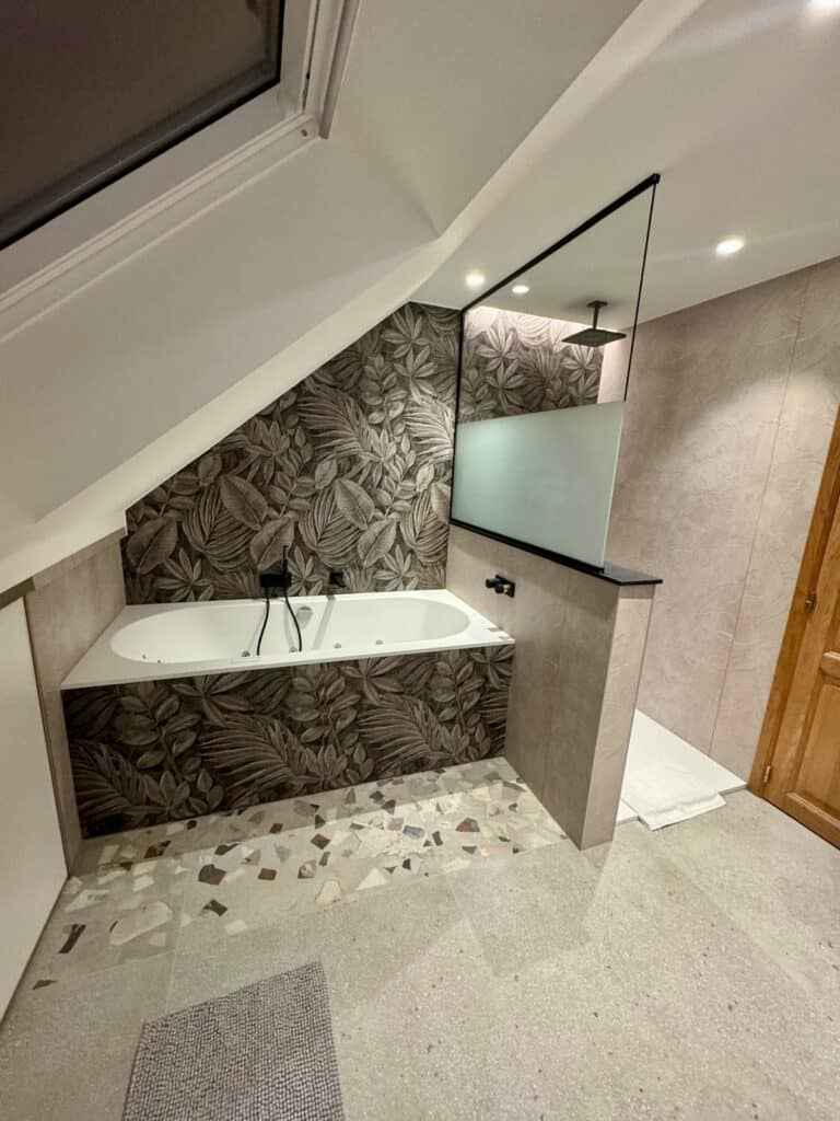 Création d’une salle de bain élégante à Verviers par Amebath : douche à l’italienne, robinetterie noire encastrée et ambiance végétale chic sous plafond mansardé.