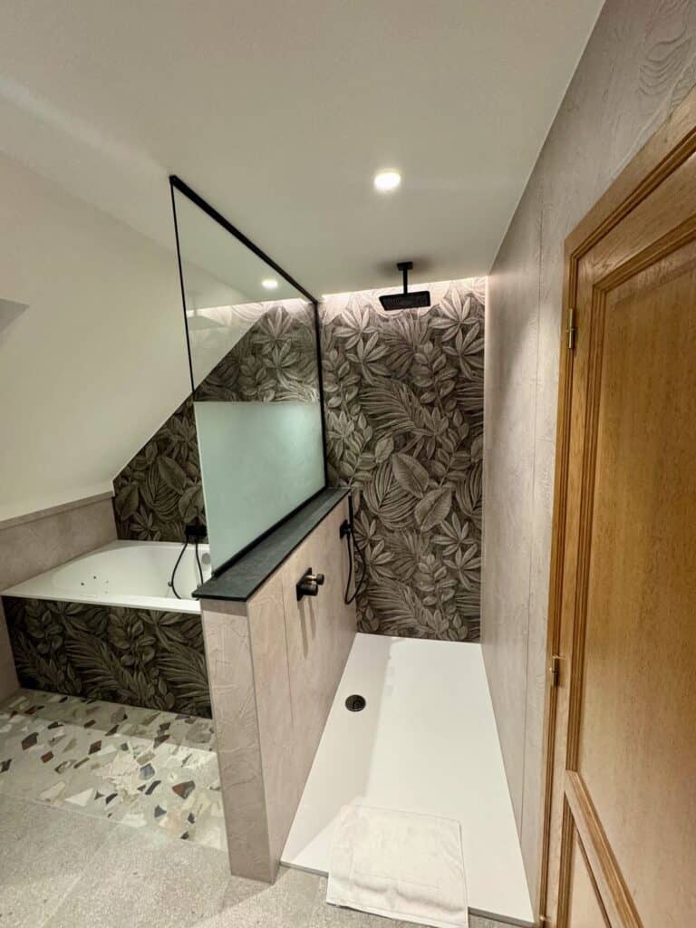 Rénovation de salle de bain sous combles à Verviers par Amebath : douche italienne avec paroi vitrée et robinetterie noire mate, murs décorés de panneaux végétaux.